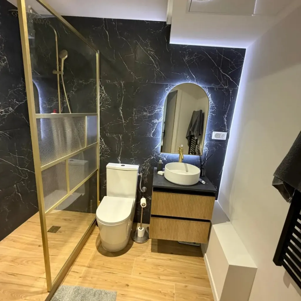 Salle de bain en grand format noir veiné, paroi de douche cadre laiton, WC, vasque ronde et miroir rétroéclairé.