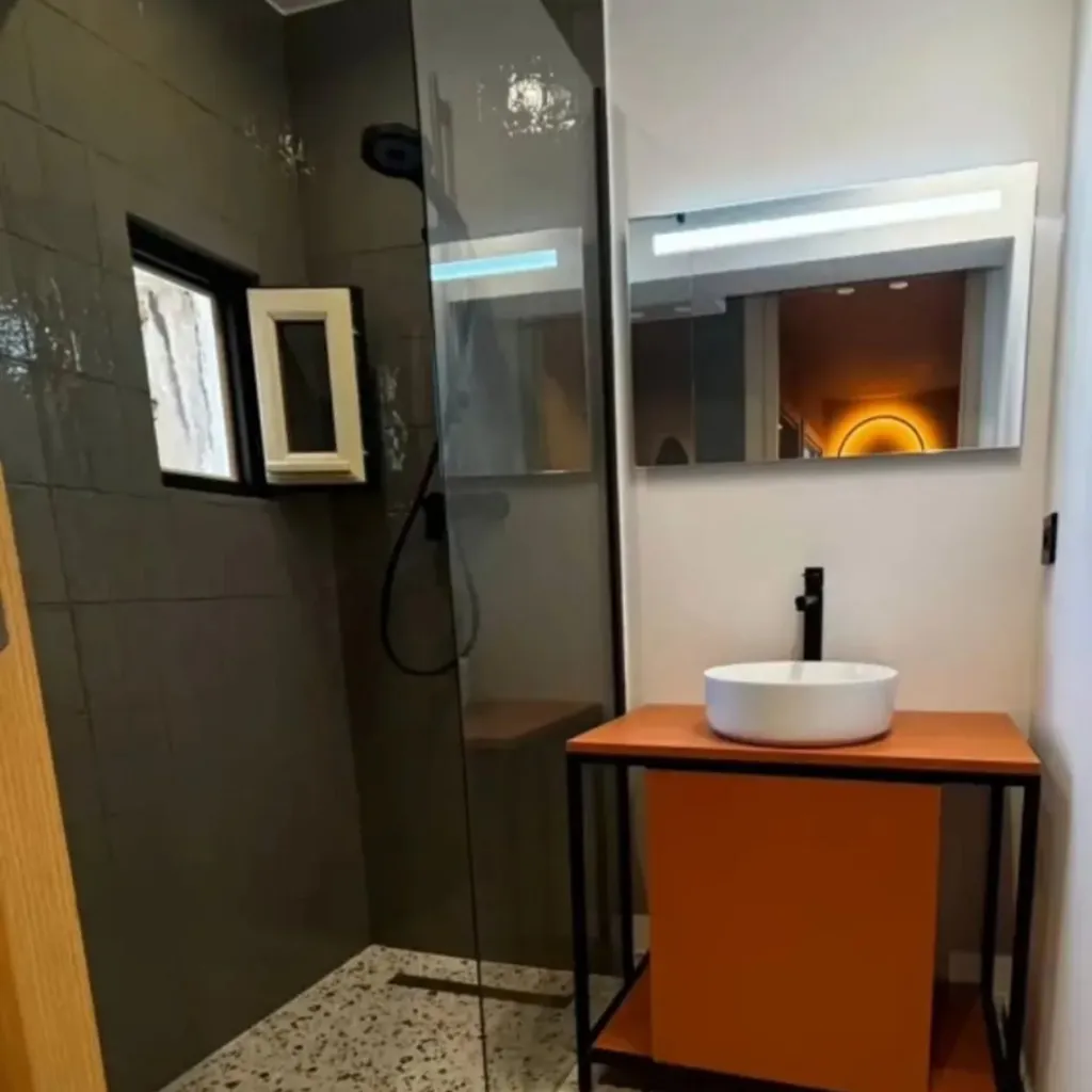Salle d'eau avec douche à l'italienne carrelée, paroi en verre, vasque sur meuble orange et miroir lumineux.