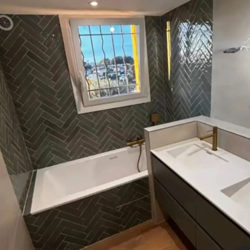 Salle de bain avec baignoire encastrée, faïence verte posée en chevrons, plan vasque blanc et robinetterie dorée.