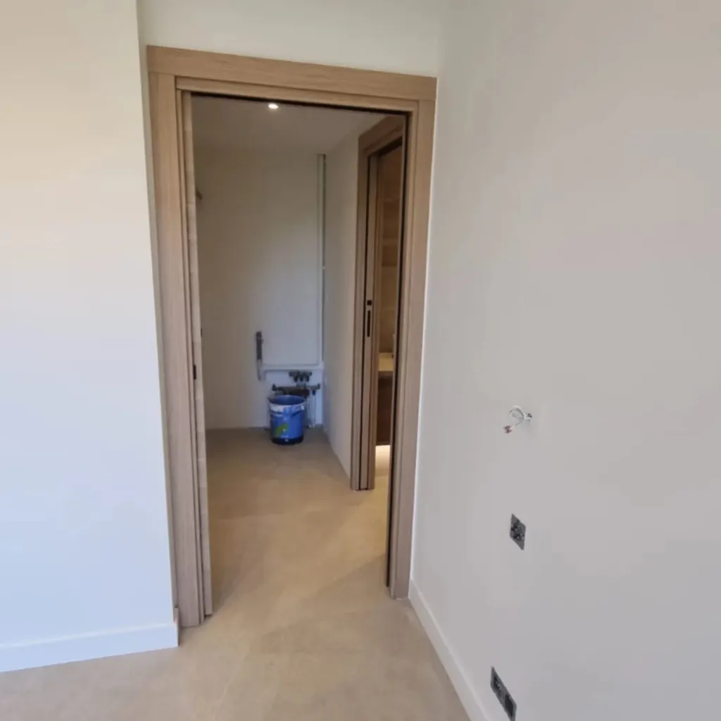 Ouverture sur une salle d'eau en cours d'installation par une porte à galandage encadrée de bois, murs lisses blancs.