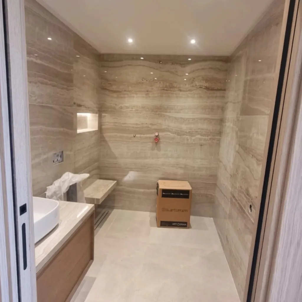 Douche de plain-pied en grand format effet travertin avec niche et banc intégrés, sol clair et vasque ronde.