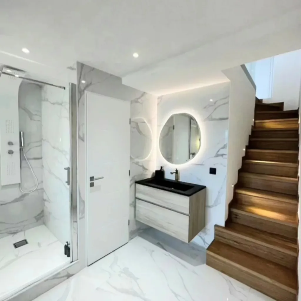 Salle de bain en carrelage effet marbre blanc avec douche vitrée, meuble vasque suspendu, miroir rétroéclairé et escalier bois.