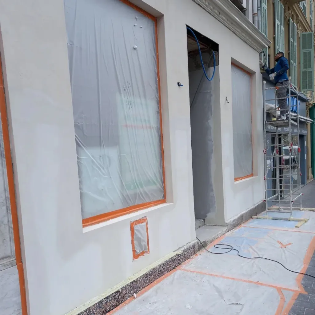 Façade de boutique enduite blanc cassé, vitrines protégées par du film plastique et échafaudage roulant avec ouvrier.