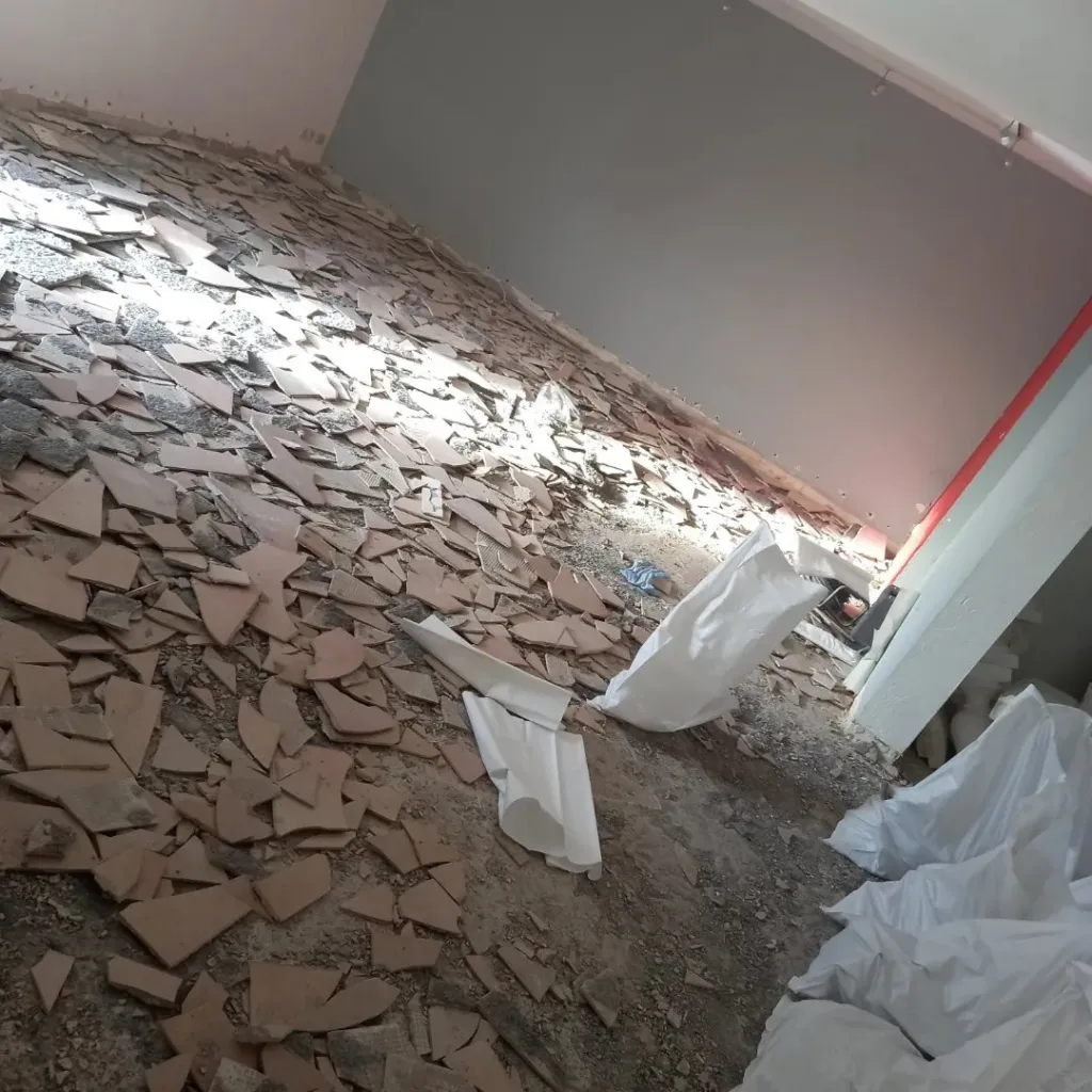 Sol d'une pièce couvert de débris de carrelage cassé après dépose, sacs de gravats au fond.