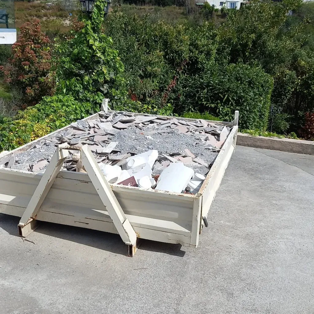 Benne en bois remplie de débris de carrelage et de gravats sur une dalle béton devant un jardin.