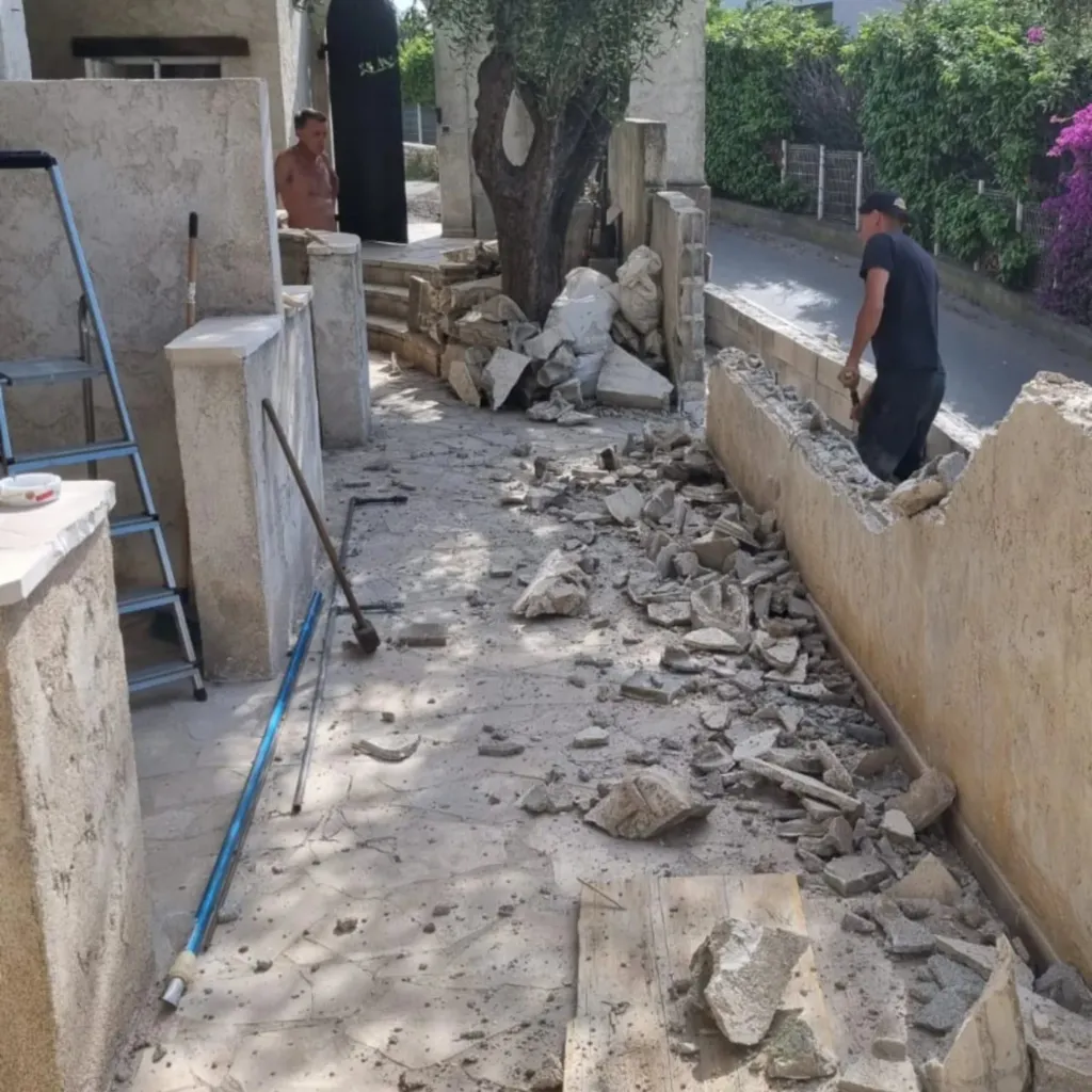 Ouvrier démolissant un muret de clôture en béton dans une allée, éclats de pierre et échelle au premier plan.