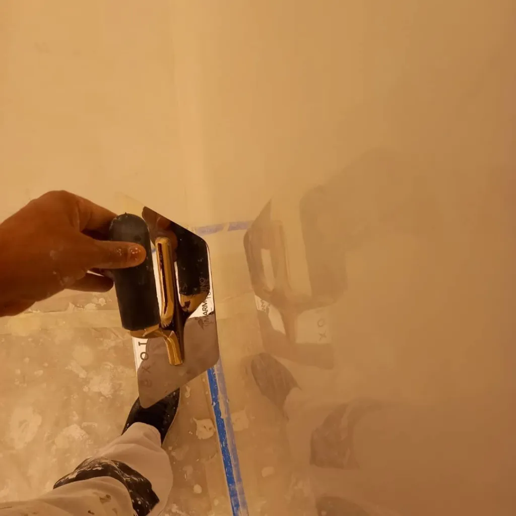 Lissage d'un enduit décoratif avec une lisseuse en acier et ruban de masquage bleu.