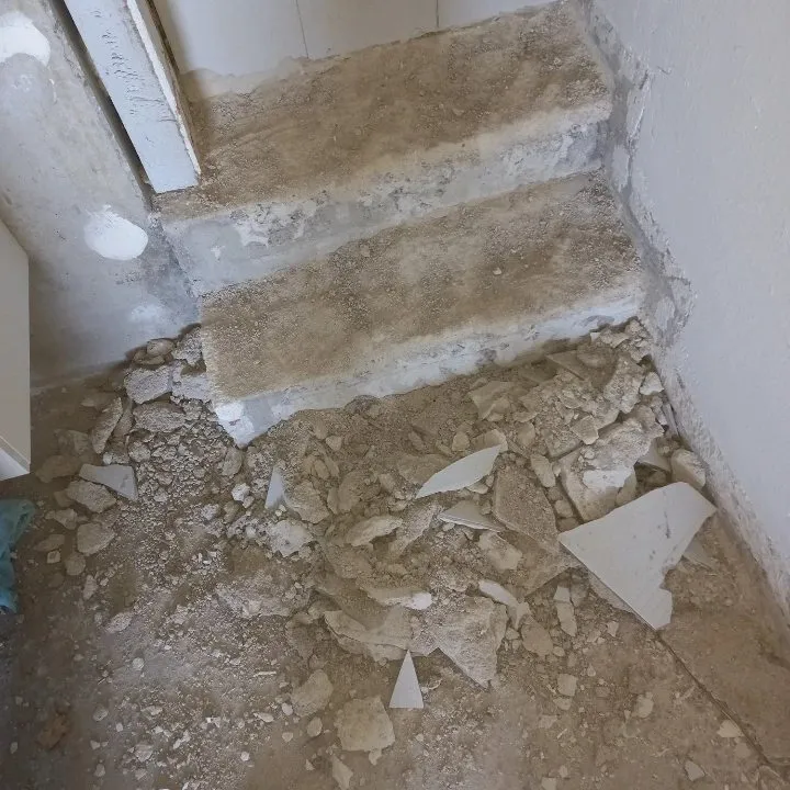 Marches d'escalier en béton décapées avec éclats de carrelage et gravats au pied.