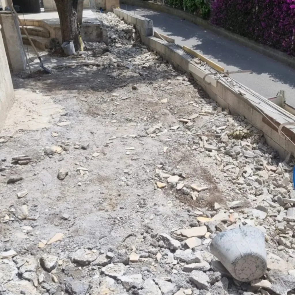 Allée dégagée jonchée de gravats avec muret en blocs à bancher le long de la route, seau renversé.