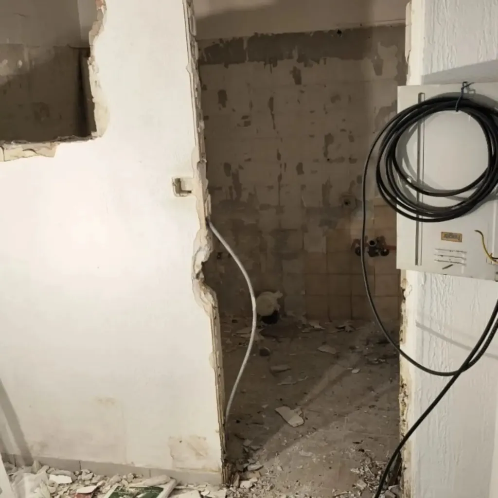 Salle d'eau dépouillée avec cloison percée, faïence arrachée et câbles électriques en attente.