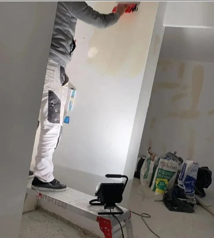 Ouvrier en tenue blanche enduisant un mur depuis un escabeau, sacs d'enduit et projecteur de chantier au sol.