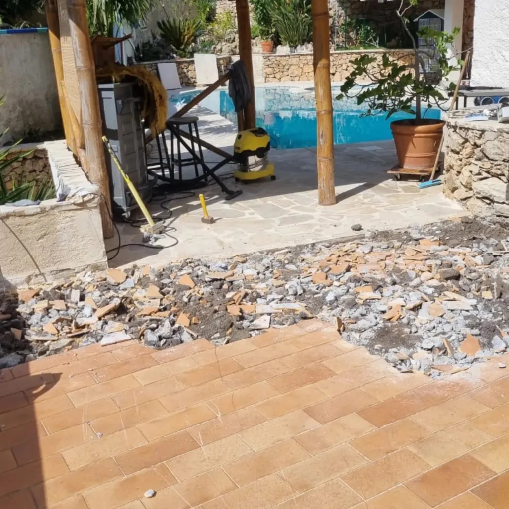 Terrasse dont le carrelage terre cuite est en cours de dépose, gravats et aspirateur de chantier près de la piscine.