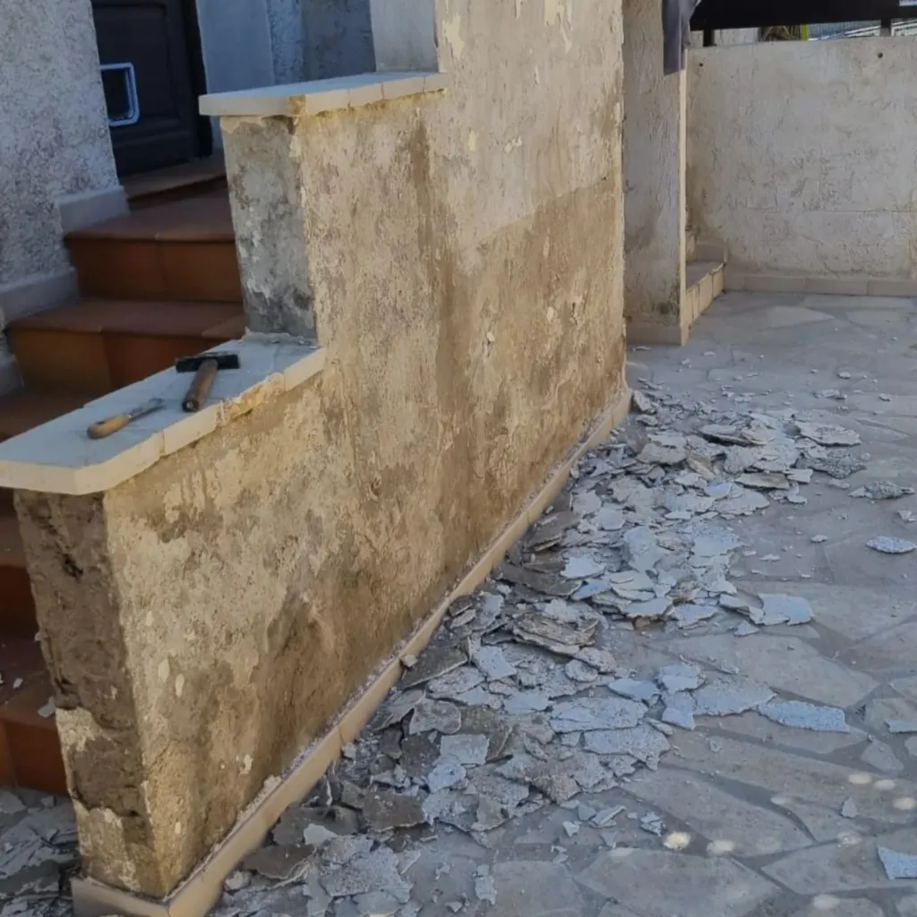 Muret d'entrée au crépi décapé, dalles de pierre irrégulières brisées au sol et escalier terre cuite à gauche.