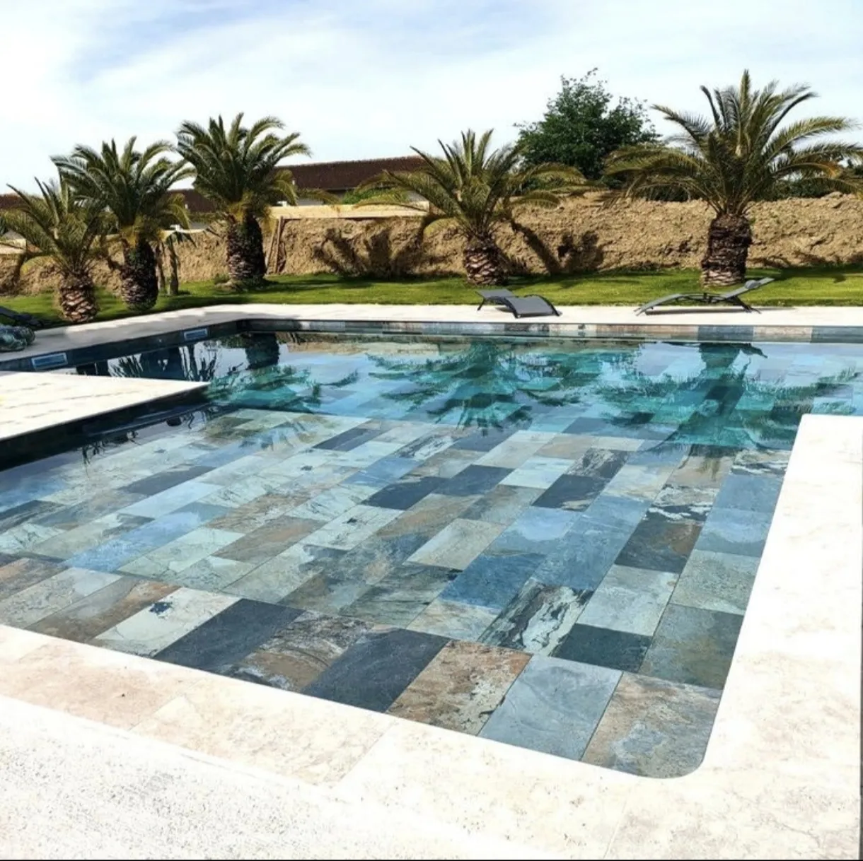 Tiled swimming pool with turquoise water surrounded by a light stone deck and lawn with palm trees