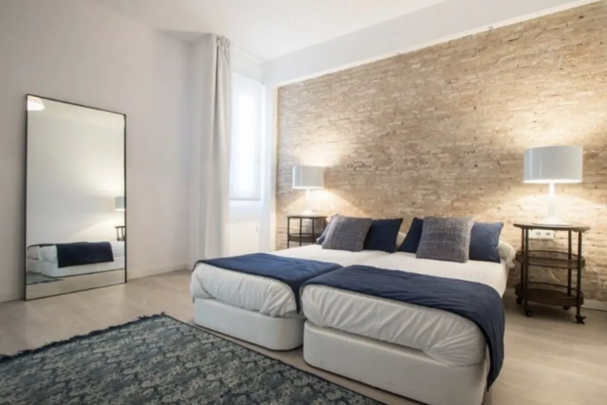 Two white beds with navy throws sit against an exposed brick wall in a bright bedroom with a tall mirror and rug